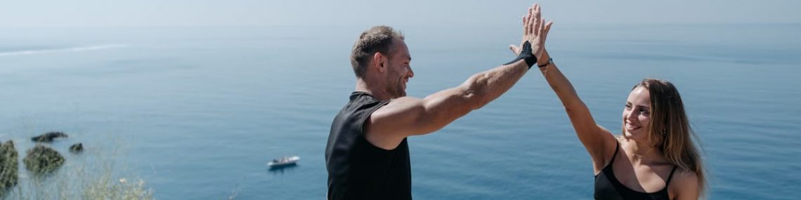 A happy couple in activewear high fives on a cliff by the ocean, enjoying a sunny day outdoors.